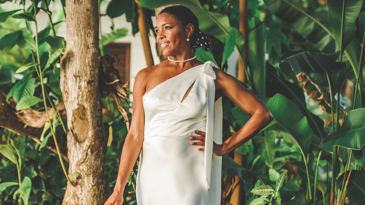 A woman in a one-shoulder white satin dress stands outdoors among tropical plants, smiling and looking to the side.